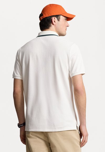Polo shirt blanc à finition texturée, avec un col vert et des fentes sur les côtés. Le modèle porte une casquette orange et un short beige.