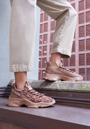 Person iført beige bukser og brune Steve Madden-sneakers, der står på udendørs trapper med en murstensvæg i baggrunden.