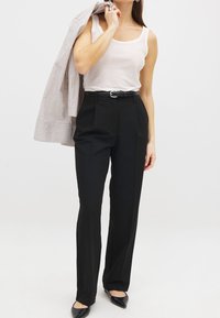 Pantalones negros ajustados de cintura alta, con pliegues y un cinturón. Combinados con una blusa sin mangas color crema y una chaqueta a rayas que se lleva sobre el hombro.