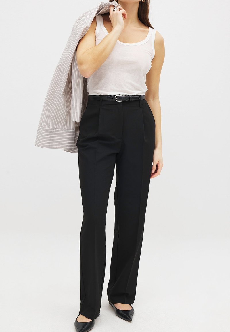 Pantalones negros ajustados de cintura alta, con pliegues y un cinturón. Combinados con una blusa sin mangas color crema y una chaqueta a rayas que se lleva sobre el hombro.