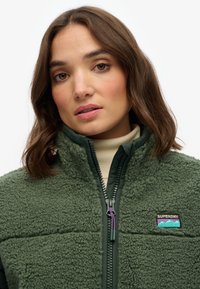 Groene fleecejas met een gestructureerd, donzig materiaal, halve rits aan de voorkant en een logopatch op de borst met paarse en turquoise accenten.