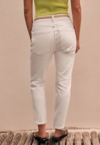White denim jeans with a fitted cut, featuring two back pockets and a mid-rise waist, paired with black slide sandals.