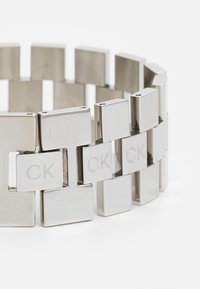 Sidabrinis metalinis apyrankės su geometriniu dizainu, kurioje yra tarpusavyje susijungę stačiakampiai grandinėlės. Išskirtinis graviravimas "CK" ant kai kurių grandinėlių.