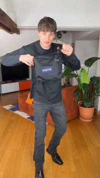 Mladi moški v črni majici in jeansa drži črno torbo Jansport čez ramo v dnevni sobi s rastlino in oranžnim kavčem.