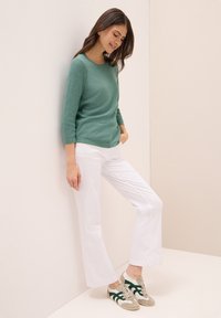 Grüner Strickpullover mit Dreiviertelärmeln, kombiniert mit weißen Schlaghosen und grau-grünen Sneakers mit grünen Akzenten. Glatte Texturen durchgehend.