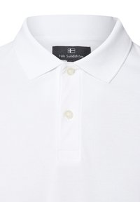 Weißes Poloshirt mit klassischem Kragen, zwei Knöpfen, strukturiertem Stoff und einem markierten schwarzen Etikett am Ausschnitt.