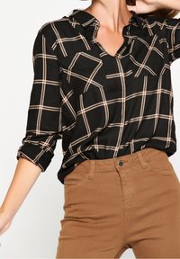 Žena oblečená v dlouhé košili s černobéžovým kostkovaným vzorem, zasunuté do hnědých kalhot s vysokým pasem, se zvednutou rukou k hlavě.