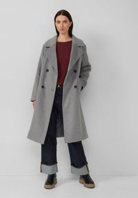 Vrouw in een lange grijze dubbelgeknoopte jas, bordeauxrode textuurtop, donkere jeans met omslag, en zwarte laarzen, staande tegen een eenvoudige muur.