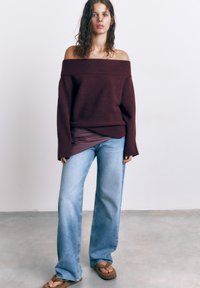 Ung kvinde med vådt hår iført off-shoulder mørkerød trøje, lyseblå brede jeans og brune klipklappere, stående på betongulv.