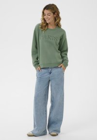 Femme souriante portant un sweat à capuche vert avec le texte "PARIS", un jean bleu clair à jambes larges, les mains dans les poches et des chaussures beige sur un fond blanc.