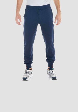 Uomo che indossa pantaloni sportivi blu navy con caviglie risvoltate e sneaker bianche con dettagli neri e arancioni, in piedi davanti a uno sfondo neutro.