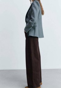 Harmaa bleiseri, jossa on jämäkät olkapäät, yhdistettynä korkeavyötäröisiin, leveälahkeisiin ruskeisiin housuihin. Molemmat on valmistettu tekstuurillisesta kankaasta.