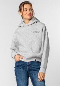 Hellgrauer Kapuzenpullover aus weichem Material, mit einer Fronttasche, Kapuze mit Zugband und gedrucktem Textdetail auf der Brust. Kombiniert mit blauen Jeans.