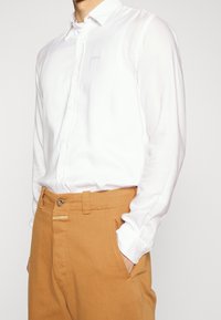 Långärmad vit skjorta med en knapp-ned krage, visad med beige byxor som har en knapp- och flikstängning, båda tillverkade av släta tyger.