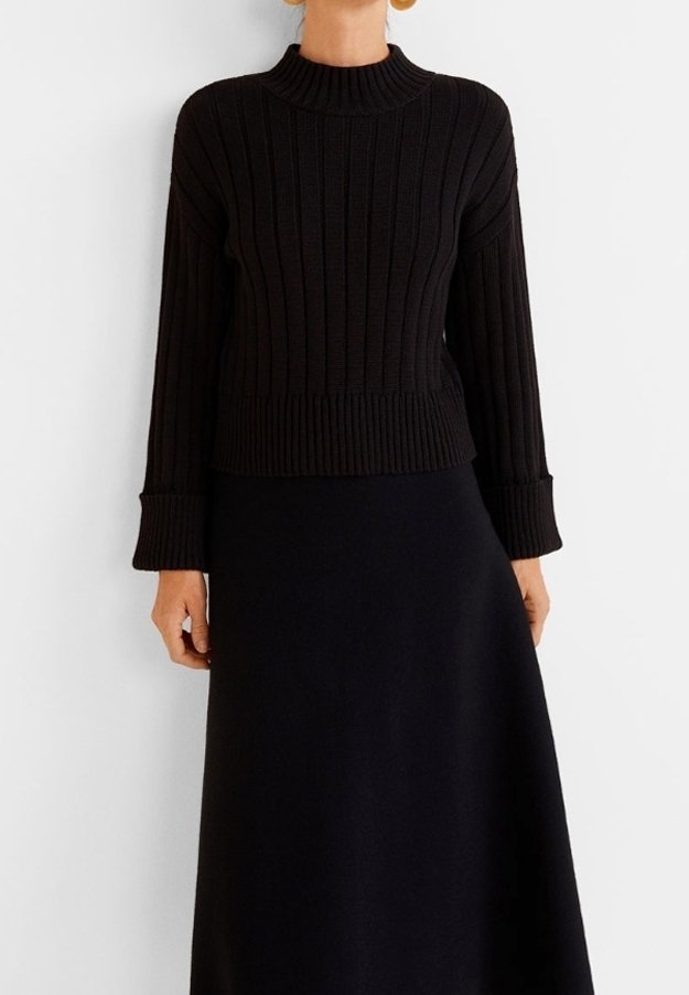 Femme portant un pull noir en tricot côtelé avec des manches longues à revers et une jupe noire évasée sur un fond blanc.