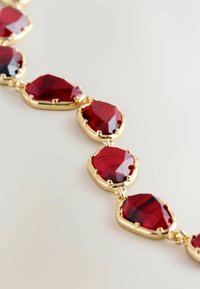 Złota bransoletka z łańcuszka z wieloma głęboko czerwonymi, fasetowanymi kamieniami połączonymi w rząd na jasnym tle.