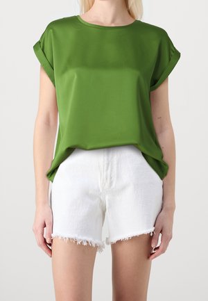 Femme portant un haut vert ample à manches courtes avec des revers roulés et un short en denim blanc effiloché, sur un fond uni.