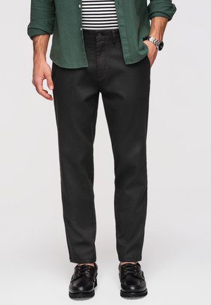 Mężczyzna ubrany w czarne spodnie, czarne skórzane buty, zieloną koszulę na czarno-białej paskowanej koszuli, z jedną ręką w kieszeni i widocznym zegarkiem na nadgarstku.