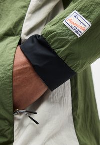 Käsi rohelise ja beeži tuulejaki taskus, must mansett, varrukal Champion Oceanside California plaastriga.