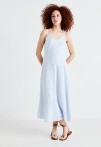 Rochie de culoare albastru deschis, până la genunchi, cu bretele subțiri, având modele brodate și detalii cu ochiuri pe întreaga suprafață. Purtați cu sandale șnururi maro.
