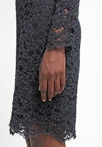 Mörkhyad hand som vilar bredvid ärmen på en knäl ång svart spetsklänning med blommotiv och vågiga kanter.