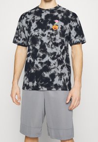 Hombre vistiendo una camiseta de tie-dye negro y gris con un pequeño logo naranja de Nike y una flor rosa en el pecho, pantalones cortos grises, de pie con los brazos relajados.
