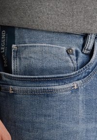 Vybledlé modré denimové džíny s viditelnou kapsou na mince, s kontrastním šitím a vytlačenou etiketou, která trčí z pásku.