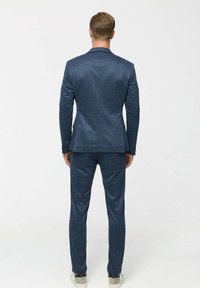 Man with short hair stands facing away wearing a fitted dark blue suit and white sneakers against a plain light background.