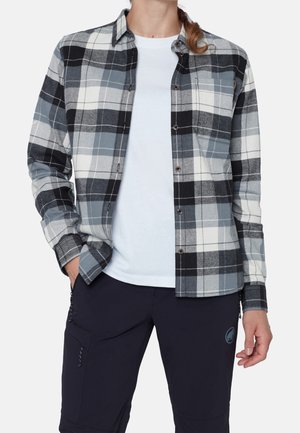 Osoba ubrana w czarno-biało-szarą kratowaną koszulę na guziki nałożoną na białą koszulkę oraz czarne spodnie z bocznymi kieszeniami.