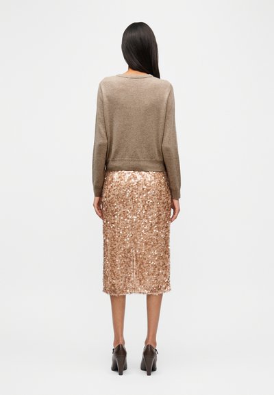 Καφέ πλεκτό πουλόβερ και φούστα μολύβι με ροζ χρυσό παγέτα που λάμπει, παρουσιάζοντας υφή ύφανσης· στυλιζαρισμένα με καφέ ψηλοτάκουνα παπούτσια.