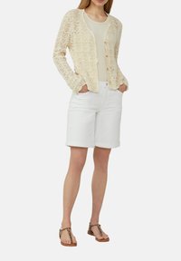 Elfenbeinfarbenes Strickcardigan mit offenem Strickdesign, kombiniert mit einem figurbetonten cremefarbenen Tanktop und weißen knielangen Shorts. Akzentuiert mit Sandalen im Leopardenmuster.
