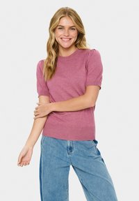 Junge Frau lächelt, trägt einen kurzärmeligen rosa Pullover und zweifarbige weite Jeans, steht mit einem Arm über dem anderen verschränkt.