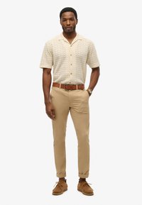 Strikket, kortærmet beige skjorte med struktureret mønster, træknapper, parret med opsmøgede, khaki slim-fit bukser og brune ruskinds sko.