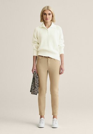 Kvinde med kort blondt hår iført hvid strikket sweater, beige bukser, hvide sneakers, der holder et foldet mørkt blomstret stof i venstre hånd.