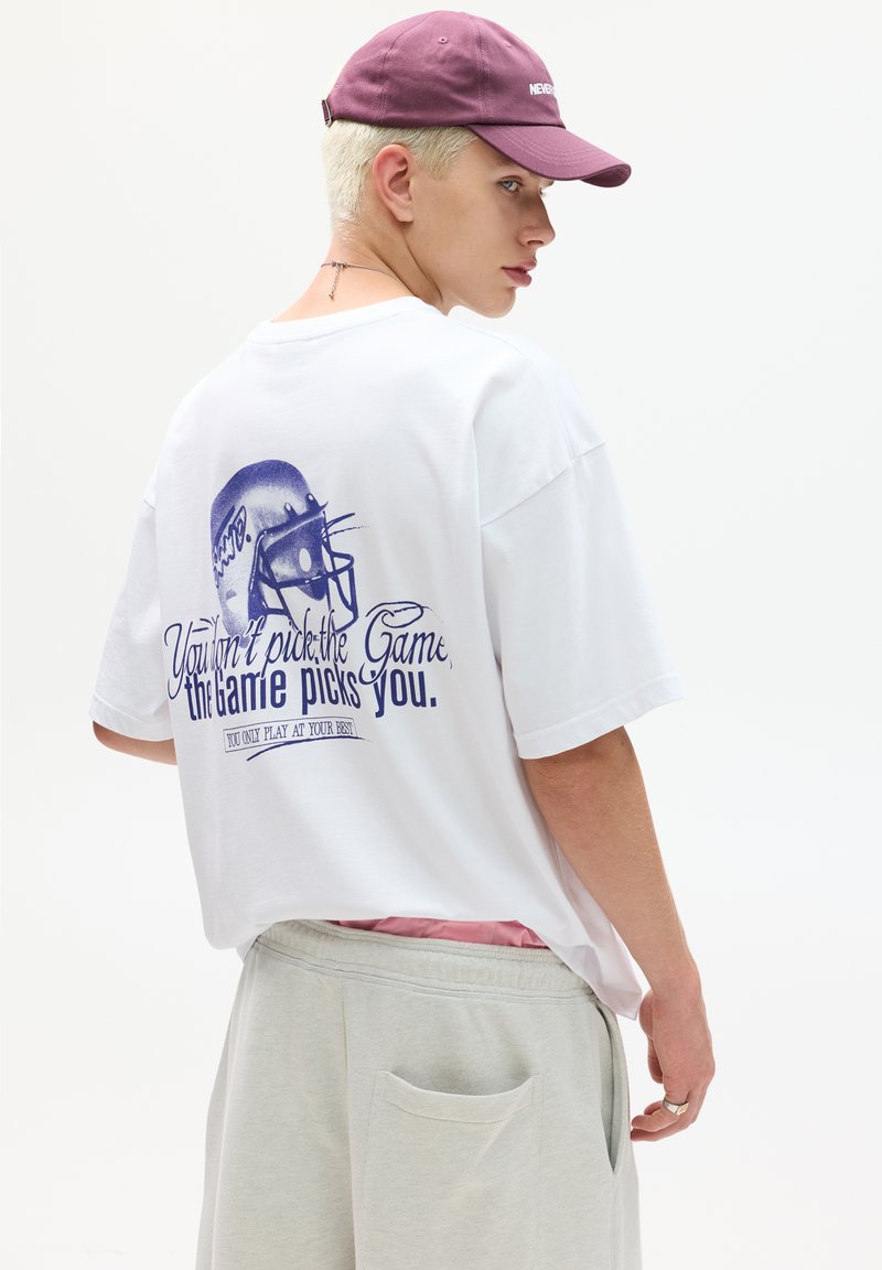 Hvid oversized t-shirt med blå grafisk af en fodboldhjelm og tekst på ryggen. Sammen med grå sweatpants og en lilla hat.