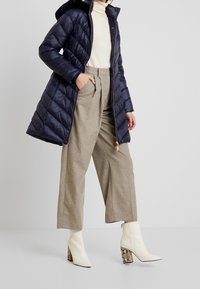 Abrigo acolchado azul marino con cierre frontal y capucha, usado sobre un suéter de tortuga color crema. Pantalones de pierna ancha en gris claro y botas blancas de tobillo con tacones de estampado de serpiente.