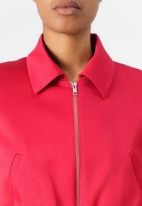 Fitted red jacket with a collared neckline, silver zipper, and side pockets. The fabric has a smooth, matte texture.