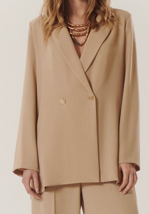 Femme portant un blazer beige croisé et un pantalon assorti avec des colliers en or superposés sur un fond neutre.