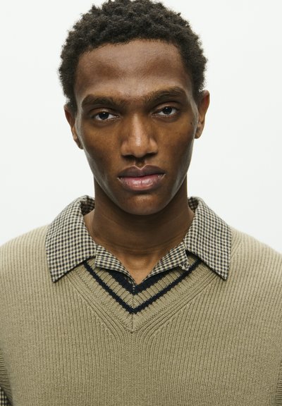 Maglione lavorato a maglia verde oliva con scollo a V, sovrapposto a una camicia a scacchi bianca e nera. Presenta una texture a costine e un colletto a contrasto.
