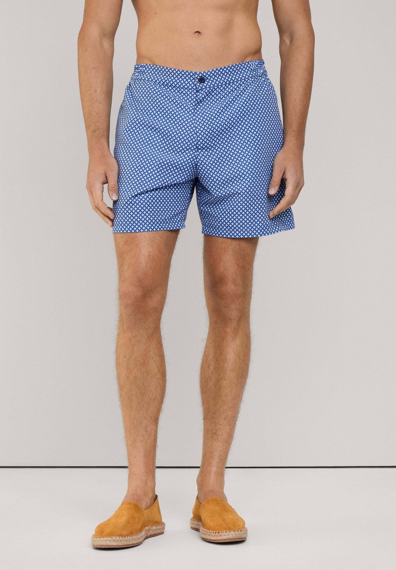 Costumi da bagno blu con un motivo geometrico, dotati di chiusura a bottone e tasche laterali, abbinati a scarpe slip-on beige.