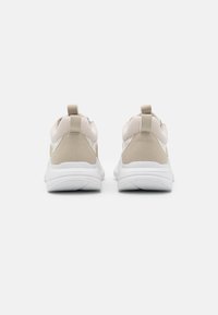 Zapatillas deportivas beige con materiales de malla y gamuza, que presentan una suela blanca, tiradores en el talón y un diseño aerodinámico.