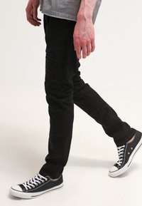 Pantalones negros ajustados con detalles acanalados en los muslos, combinados con zapatillas negras y una camisa gris, en un fondo neutral.