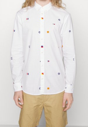 Camisa blanca de manga larga con botones y pequeños patrones coloridos de banderas náuticas, combinada con pantalones cortos de color marrón claro.