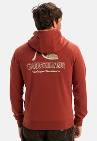 Sweat à capuche rouge avec un grand logo graphique blanc au dos, présentant une capuche à cordon de serrage et des poignets côtelés. Texture douce, design décontracté.