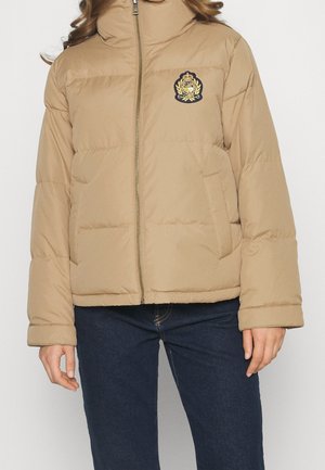 Beżowa pikowana kurtka z przednim zamkiem, stojącym kołnierzem, bocznymi kieszeniami oraz wyszywanym herbem na lewej piersi, noszona z ciemnoniebieskimi dżinsami.