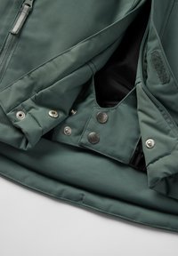 Nærbillede af en mørkegrøn jakke, der viser metaltrykknapper, lynlås, velcro-patch og overlapninger af stoflag på en lys baggrund.