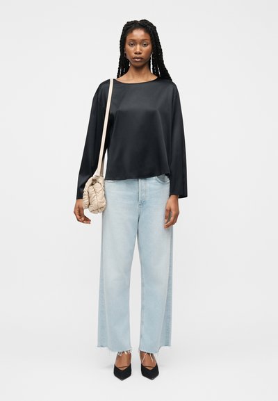 Γυναίκα που στέκεται φορώντας μαύρο μακρυμάνικο τοπ, ανοιχτού μπλε τζιν με φαρδιά πόδια και ξέφτια στα τελειώματα, μαύρα μυτερά παπούτσια, και κρατώντας μπεζ καπιτονέ τσάντα ώμου.