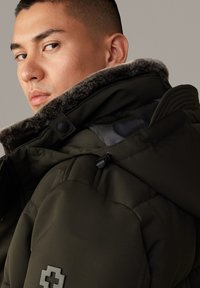 Olivengroene pufferjack met een kraag van fauxbont, voorzien van zwarte hardware en gestikte details. Close-up van de stofstructuur en het design.