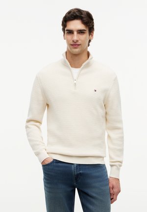 Nuori mies pukeutuneena kermaiseen, puolivetoketjulliseen neulepaitaan, jossa on logo rinnassa, kädet taskuissa ja siniset farkut, seisoo valkoista taustaa vasten.