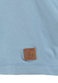 Hellblaues Gewebe mit einem braunen quadratischen Lederetikett, das nahe dem Saum eingenäht ist, geprägt mit "KEY LARGO" und einem kreisförmigen Logo.